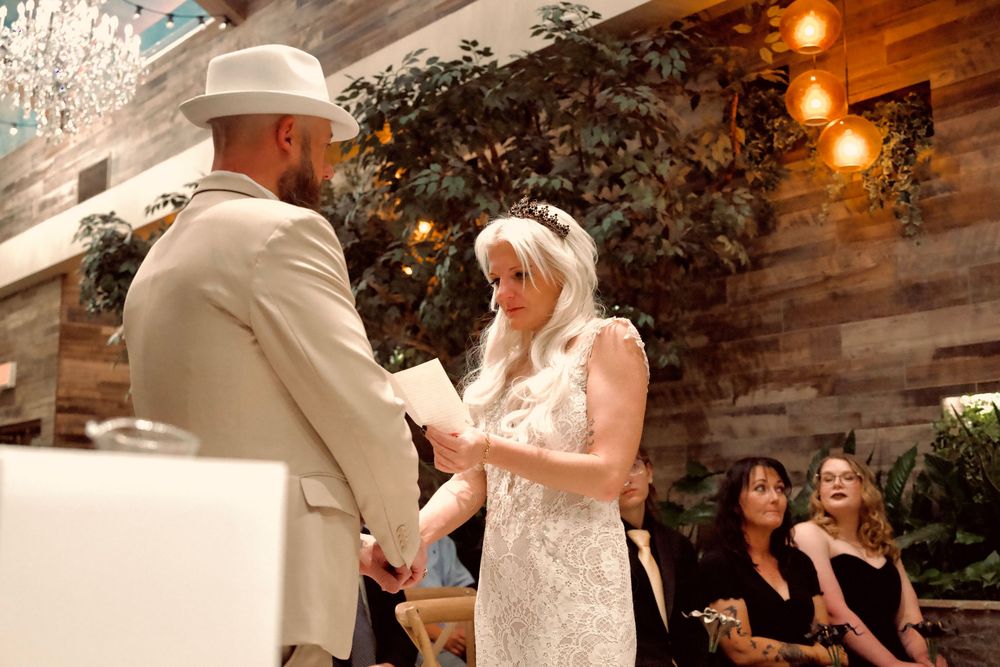 Bride reads vows to groom in white hat during indoor wedding ceremony with warm string lights and greenery.