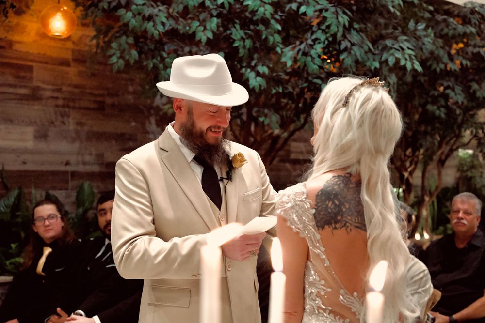 Groom in white hat and cream suit places ring on brides finger during indoor wedding ceremony.