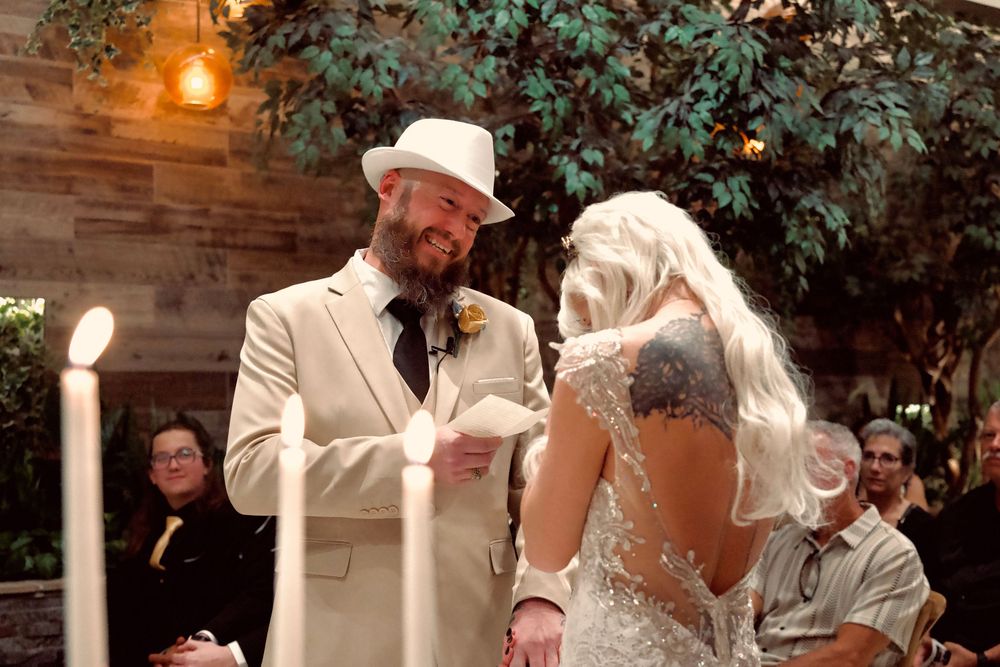 Groom in white hat and tan suit reads vows to blonde bride in lace wedding dress during indoor ceremony.