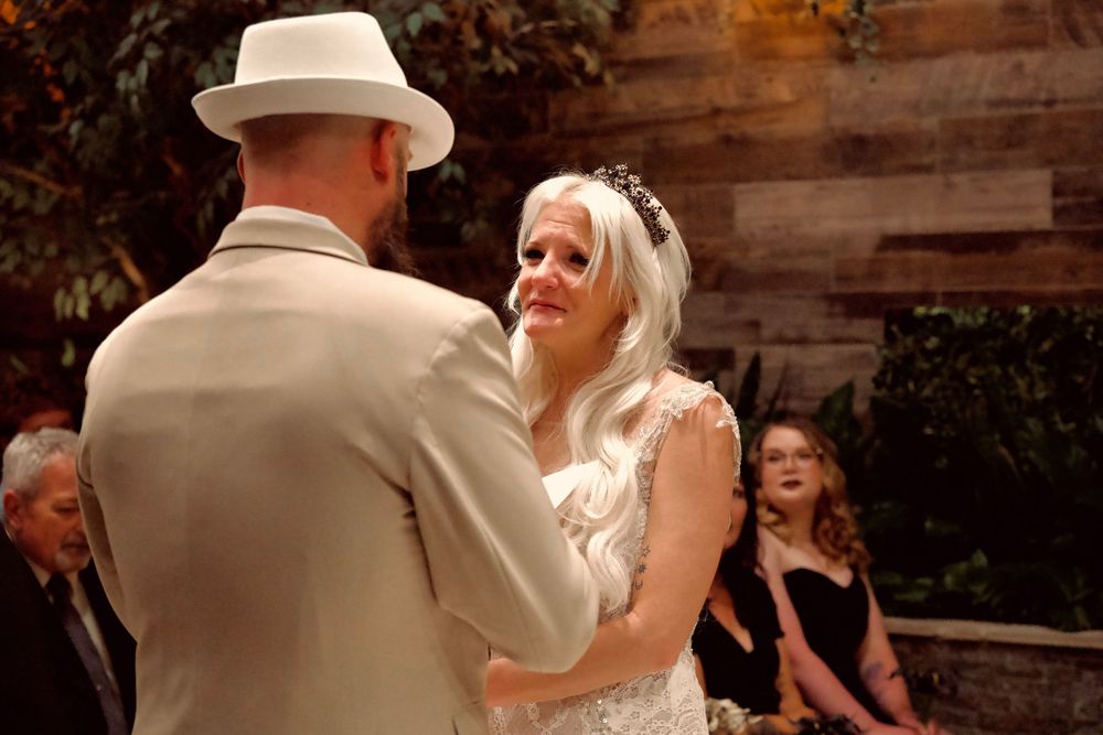 Emotional bride in white lace gown and tiara crying during wedding ceremony while holding groom's hands.