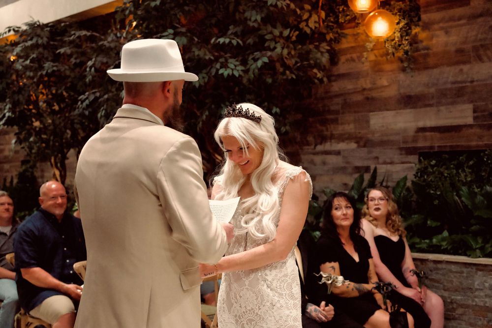 Blonde bride in lace gown smiles while holding hands with groom in cream suit at wedding ceremony.