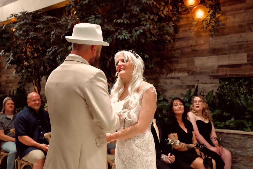 Bride in lace gown smiles joyfully at groom in white fedora during wedding ceremony with guests seated behind.