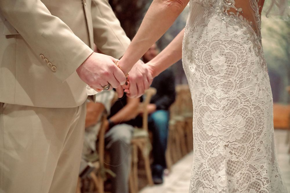 Close-up of bride and groom holding hands during wedding ceremony, bride in lace gown.