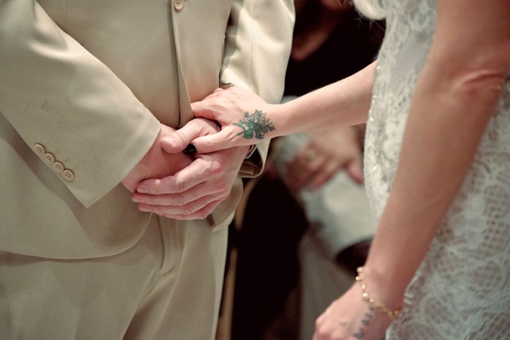 Bride and groom holding hands during wedding ceremony, groom in beige suit, bride with floral wrist tattoo.