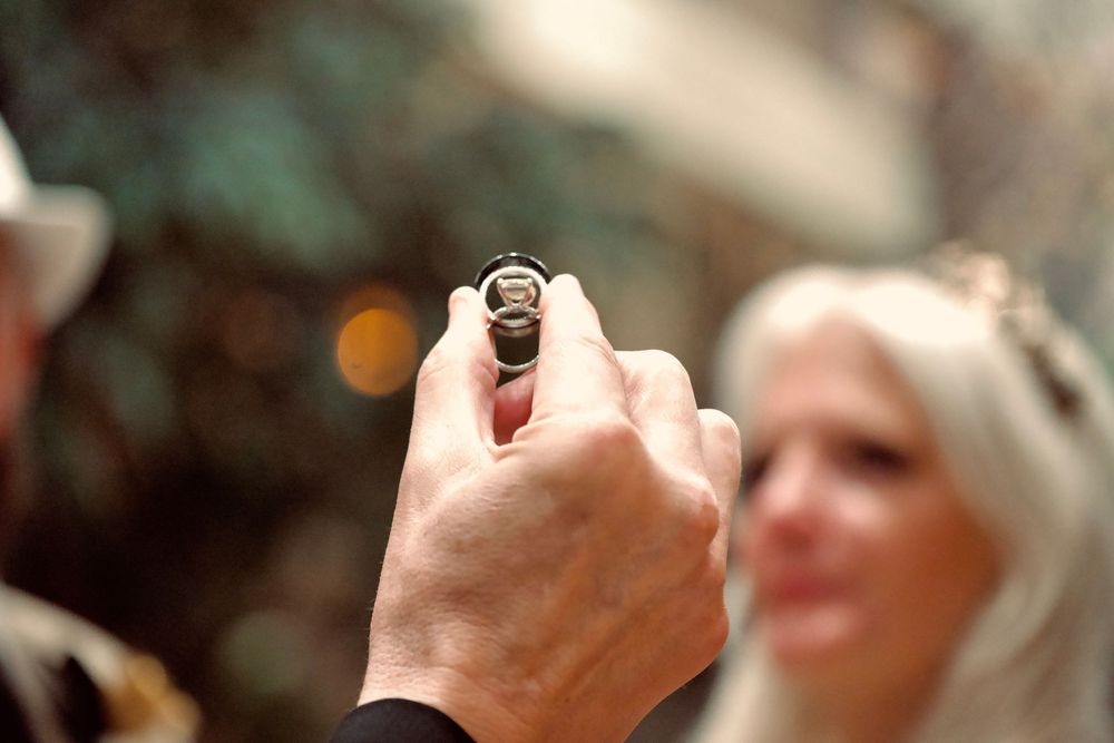 Officiant holds up wedding ring during outdoor ceremony with bride visible in background.