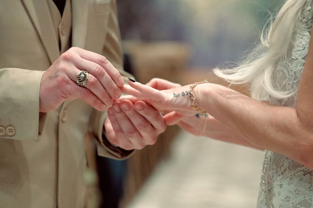 Groom placing wedding ring on brides finger during ceremony, close-up of hands with tattoos.