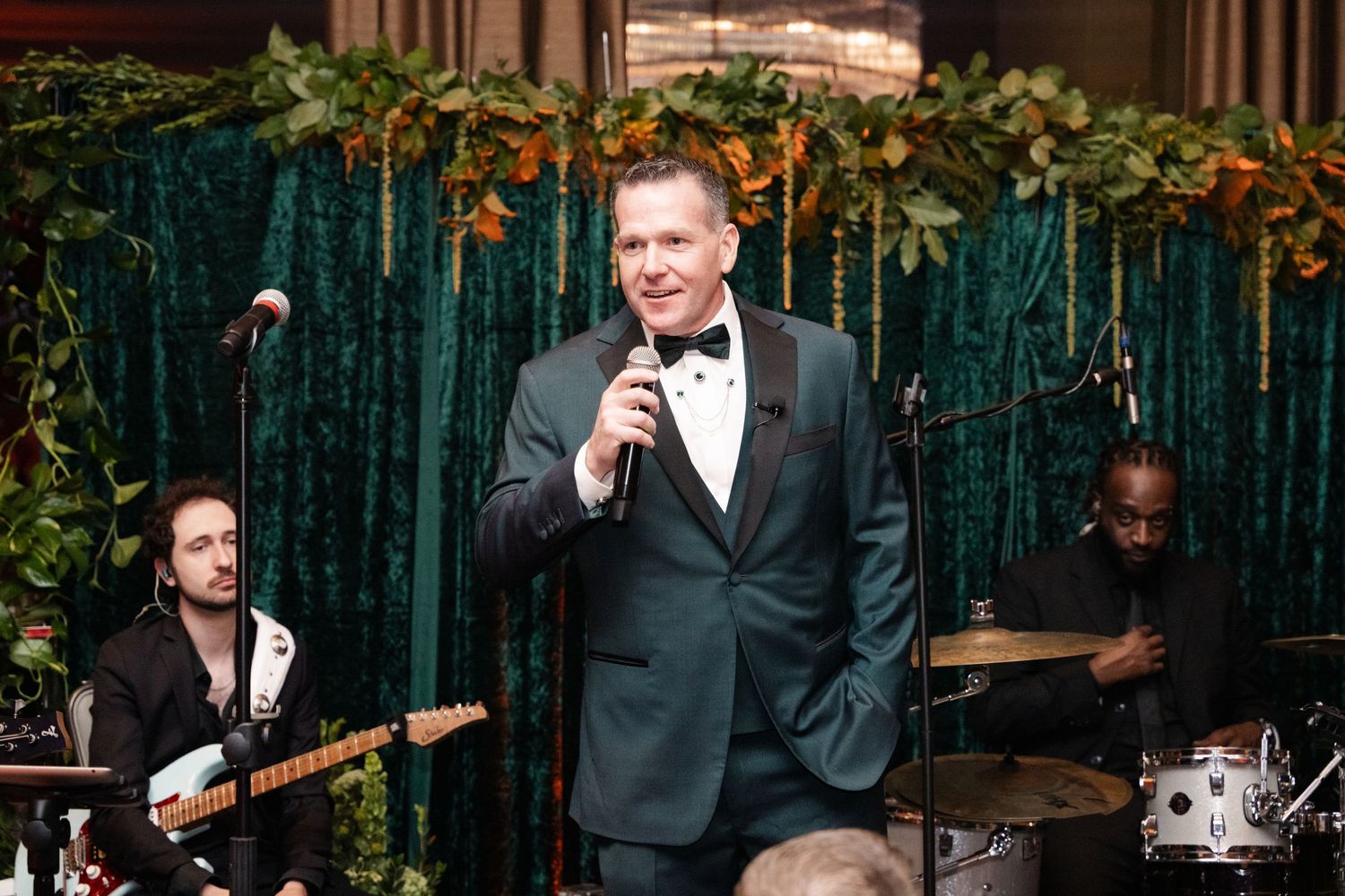 Man in green tuxedo speaks into microphone at elegant event with live band and lush green velvet backdrop.