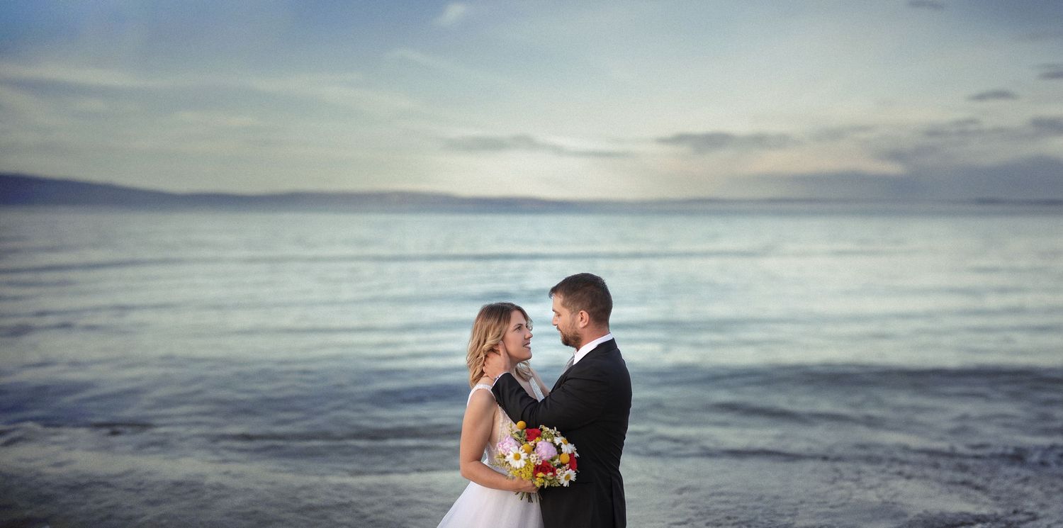 Wide-angle cinematic wedding photography of a bride and groom embracing by the calm sea under a cloudy sky.