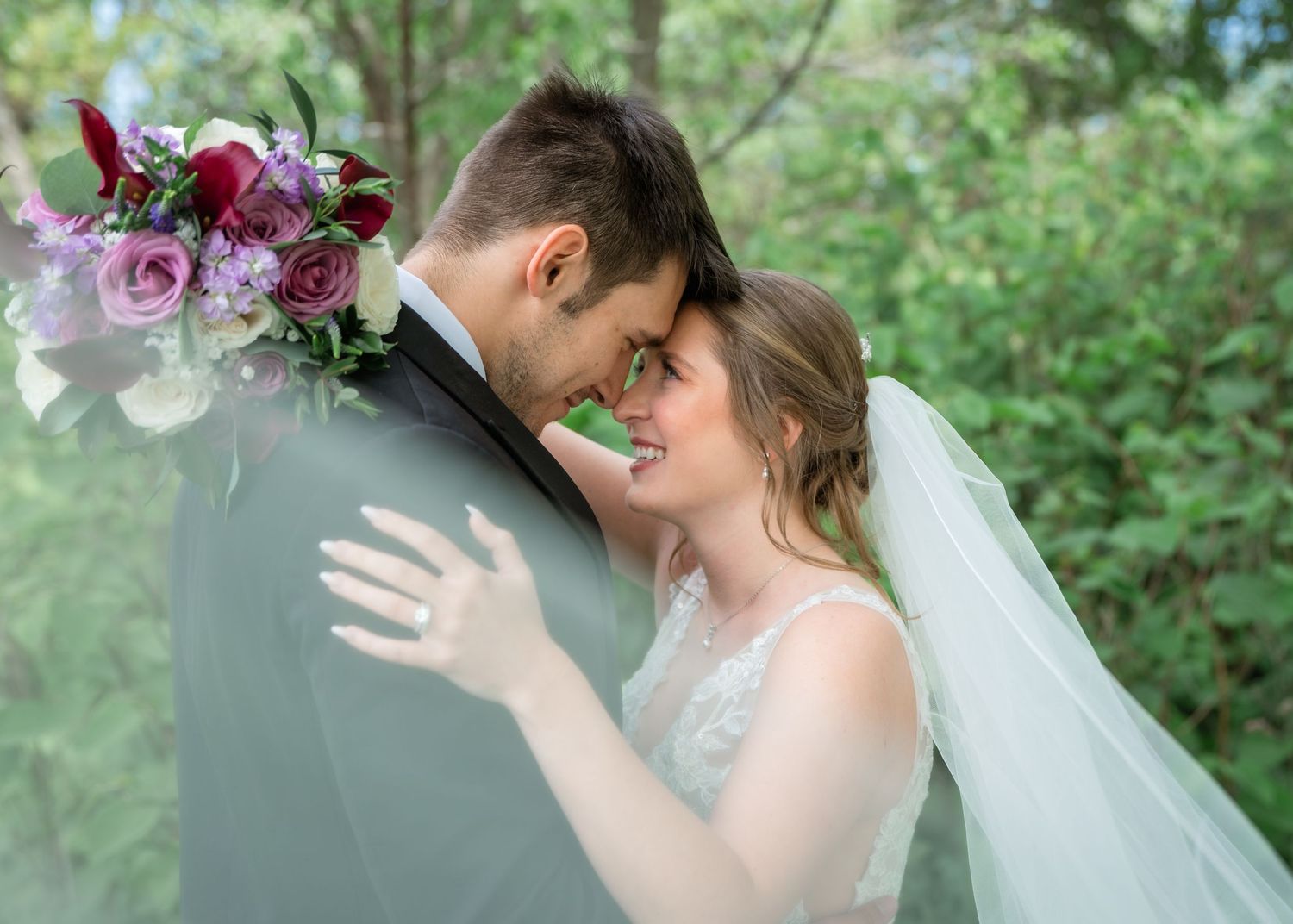 A bride and groom share a romantic forehead touch, surrounded by lush greenery, as she holds a bouquet of red and pink flowers.
