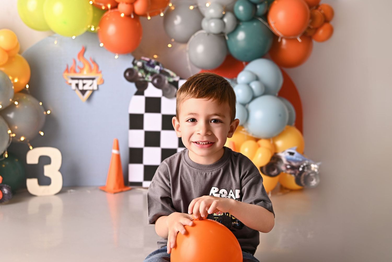 Toddler boy sits in wooden chair at Cars-themed 2nd birthday cake smash with balloons and Route 66 decorations.