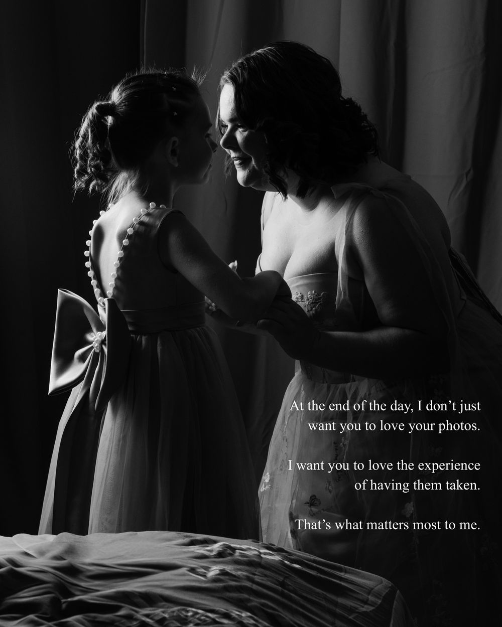 A bride shares a tender black and white moment with a young flower girl in soft window light.