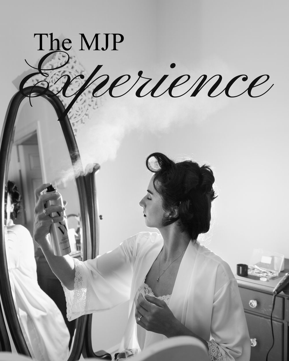 A bride in a white robe sprays perfume while gazing into an ornate mirror during wedding preparations.