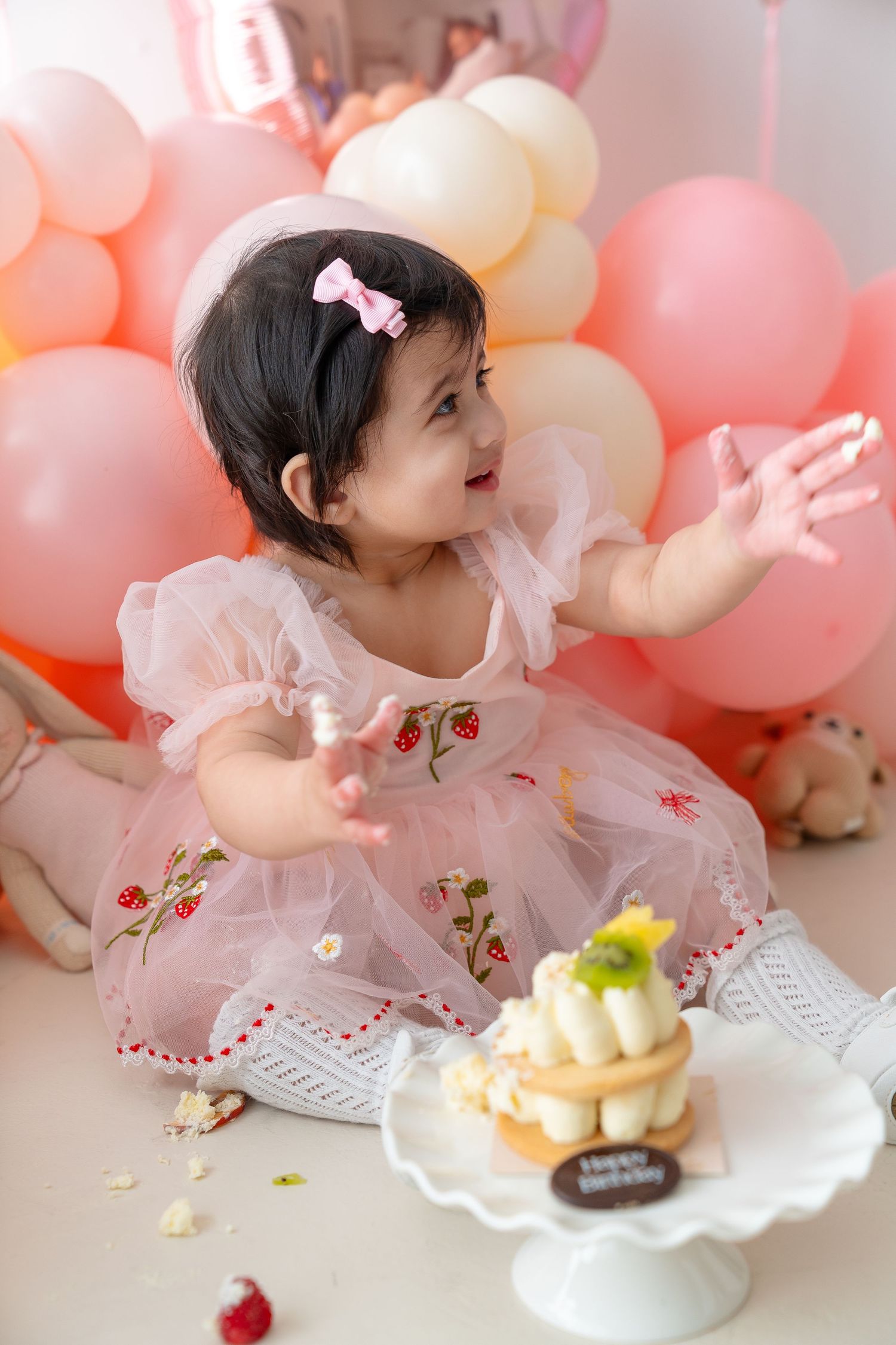 Baby girl in pink dress reaches for birthday cake surrounded by pastel balloons at first birthday cake smash.