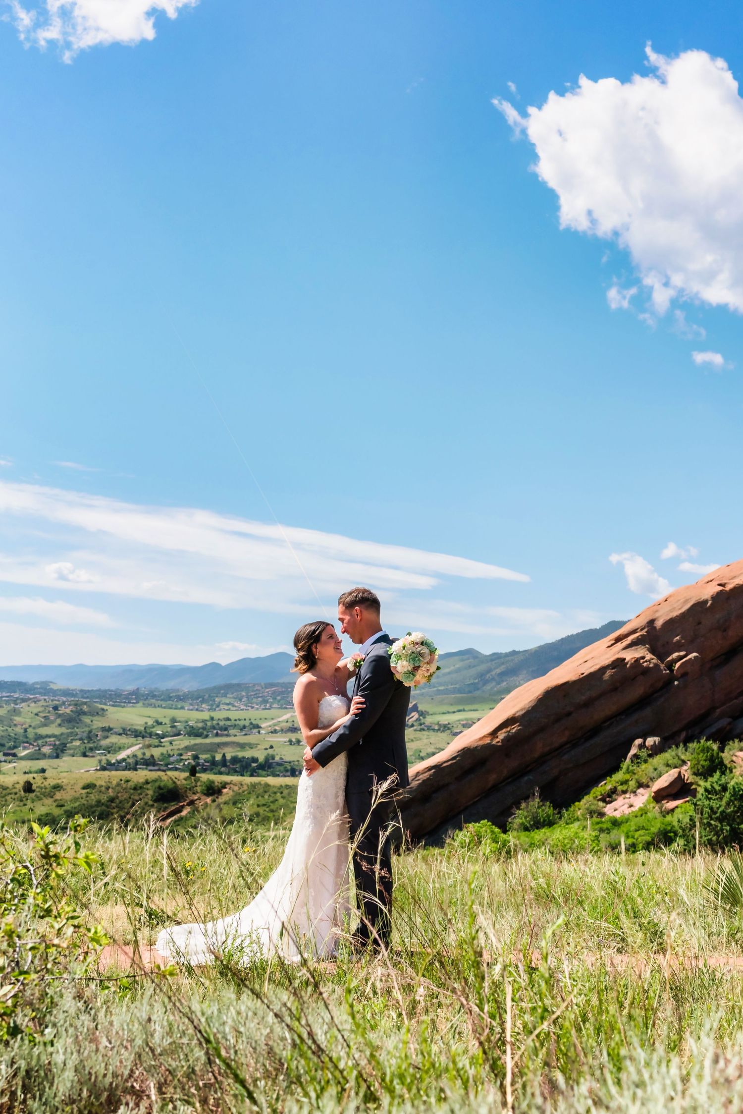 Bride and groom embrace at Red Rocks Colorado with stunning mountain valley views under a bright blue sky.