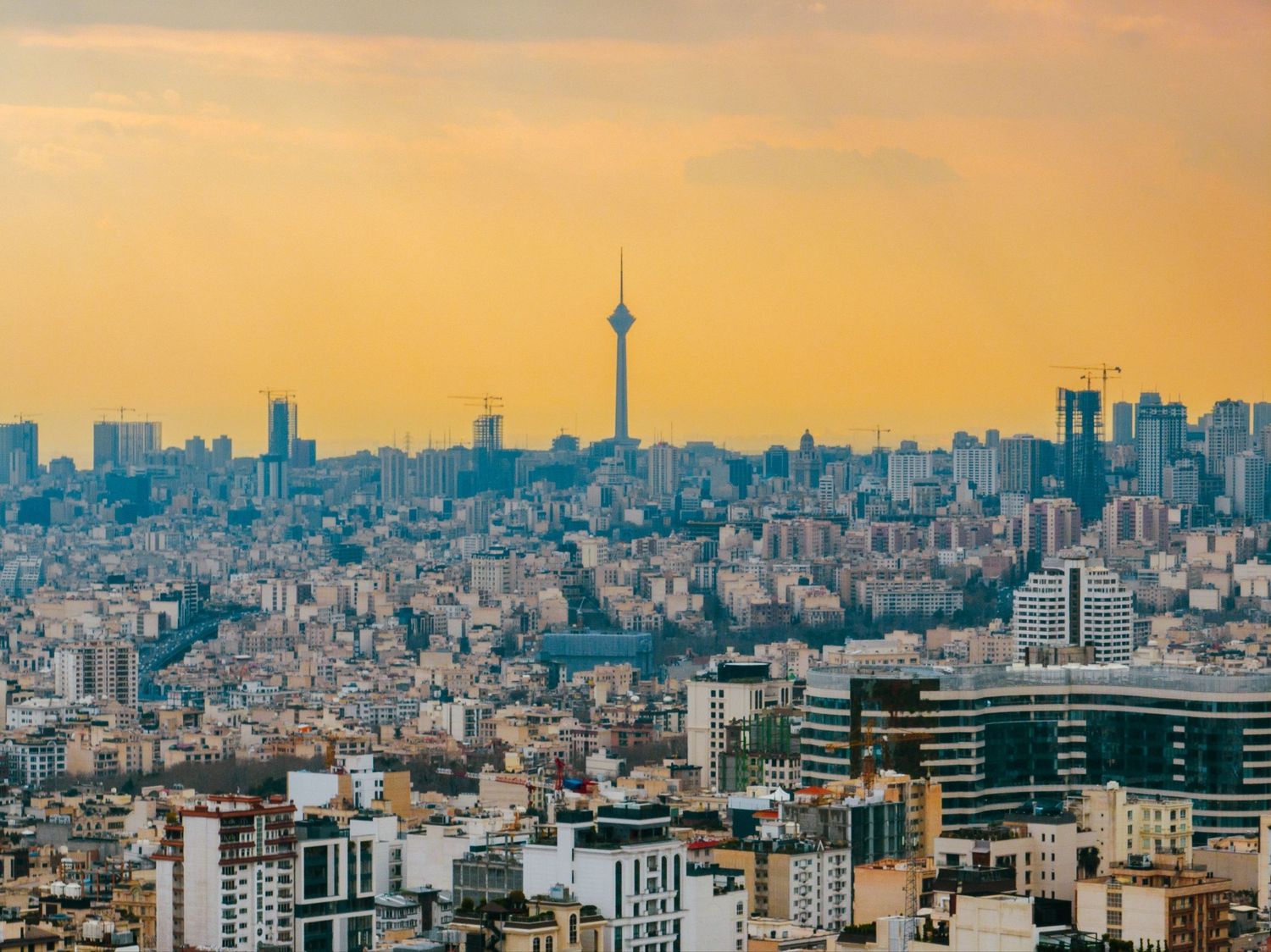 Sprawling cityscape of Tehran, Iran, at sunset, with the tall Milad Tower silhouetted against an orange sky above dense rows of apartment bu