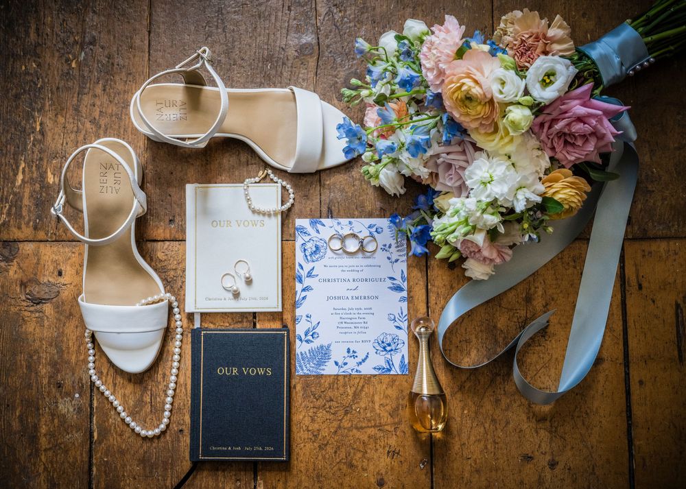 Wedding flat lay with bridal heels, bouquet, rings, vow books, and floral invitation on wood floor.