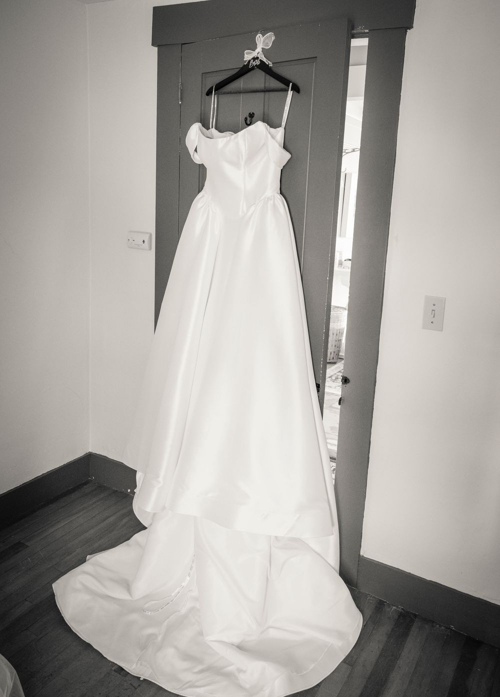 Elegant white off-shoulder wedding gown with cathedral train hanging on door in black and white photo.