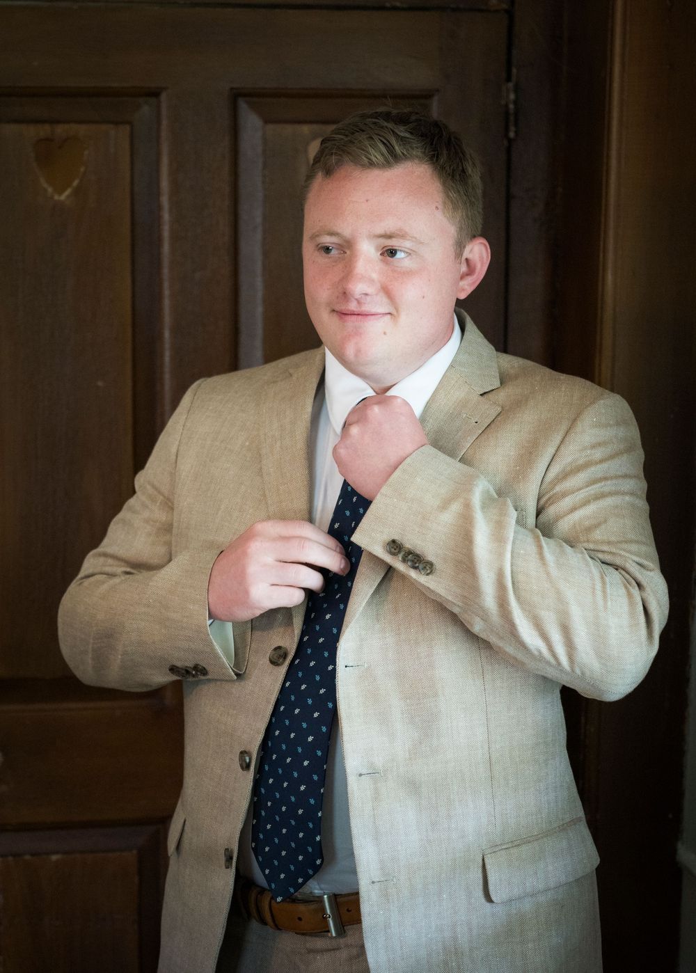 Groom in tan suit adjusting navy polka dot tie before wedding ceremony.