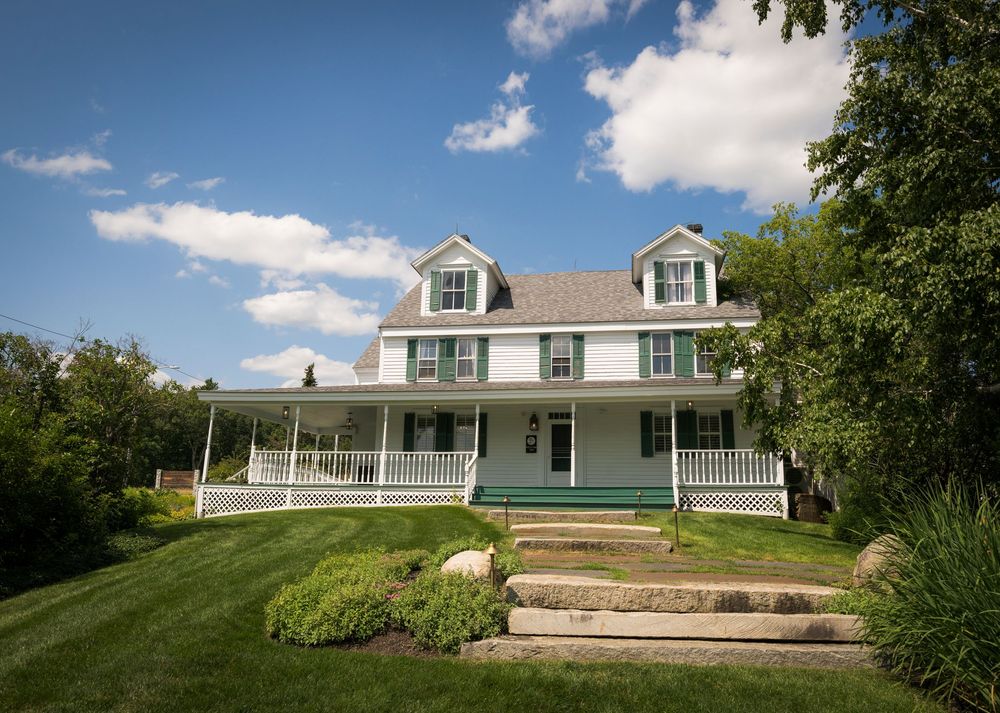 Charming white farmhouse with green shutters and wrap-around porch surrounded by lush greenery.