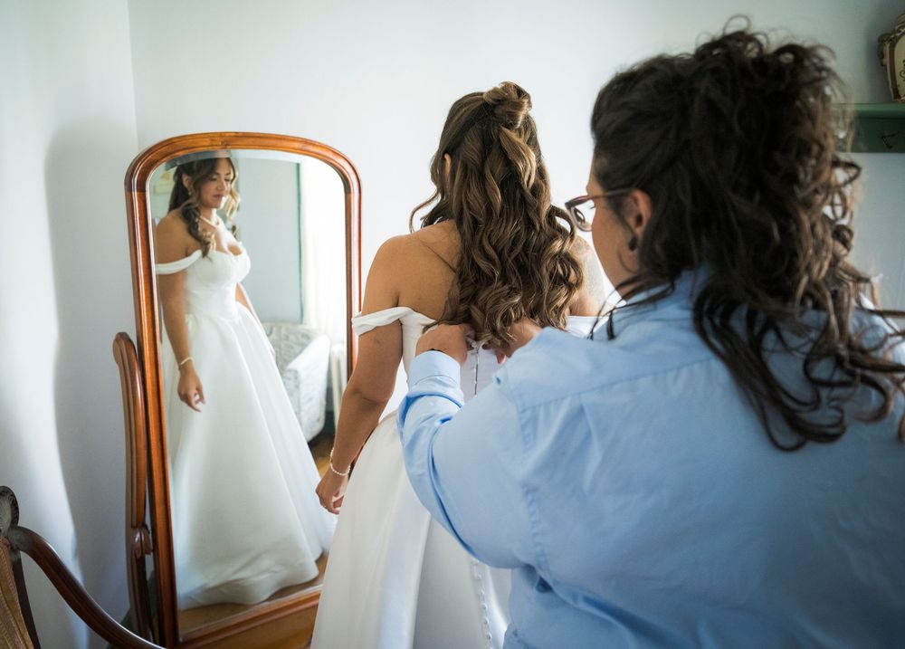 Bride in white wedding gown getting dress fastened by helper while viewing reflection in elegant wooden mirror.