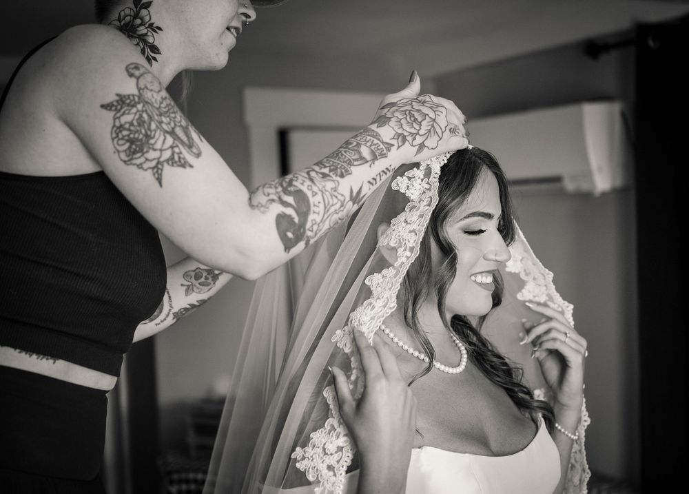 Black and white photo of bride smiling as bridesmaid adjusts her lace wedding veil.
