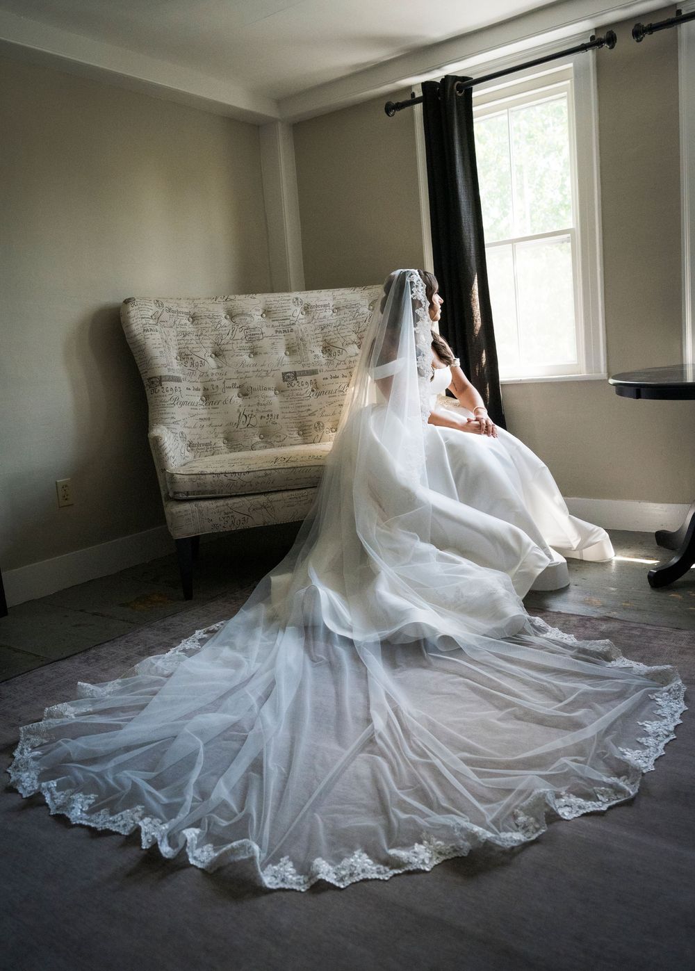 Bride in a white wedding gown with a long lace-trimmed cathedral veil sitting by a window in a sunlit room.