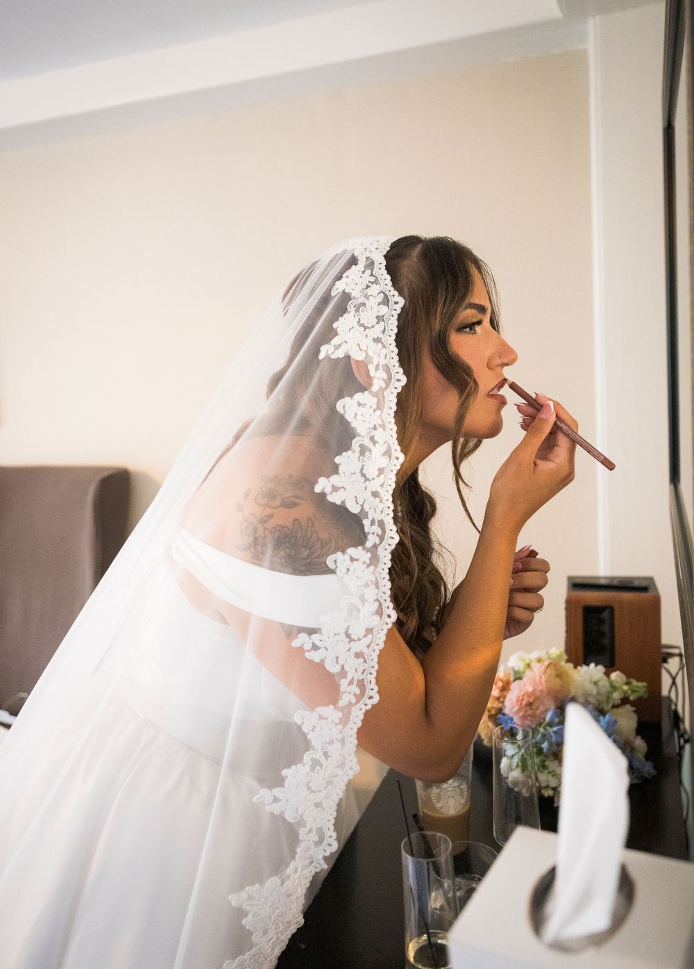 Bride in lace-trimmed veil applies lipstick at mirror while getting ready for her wedding ceremony.