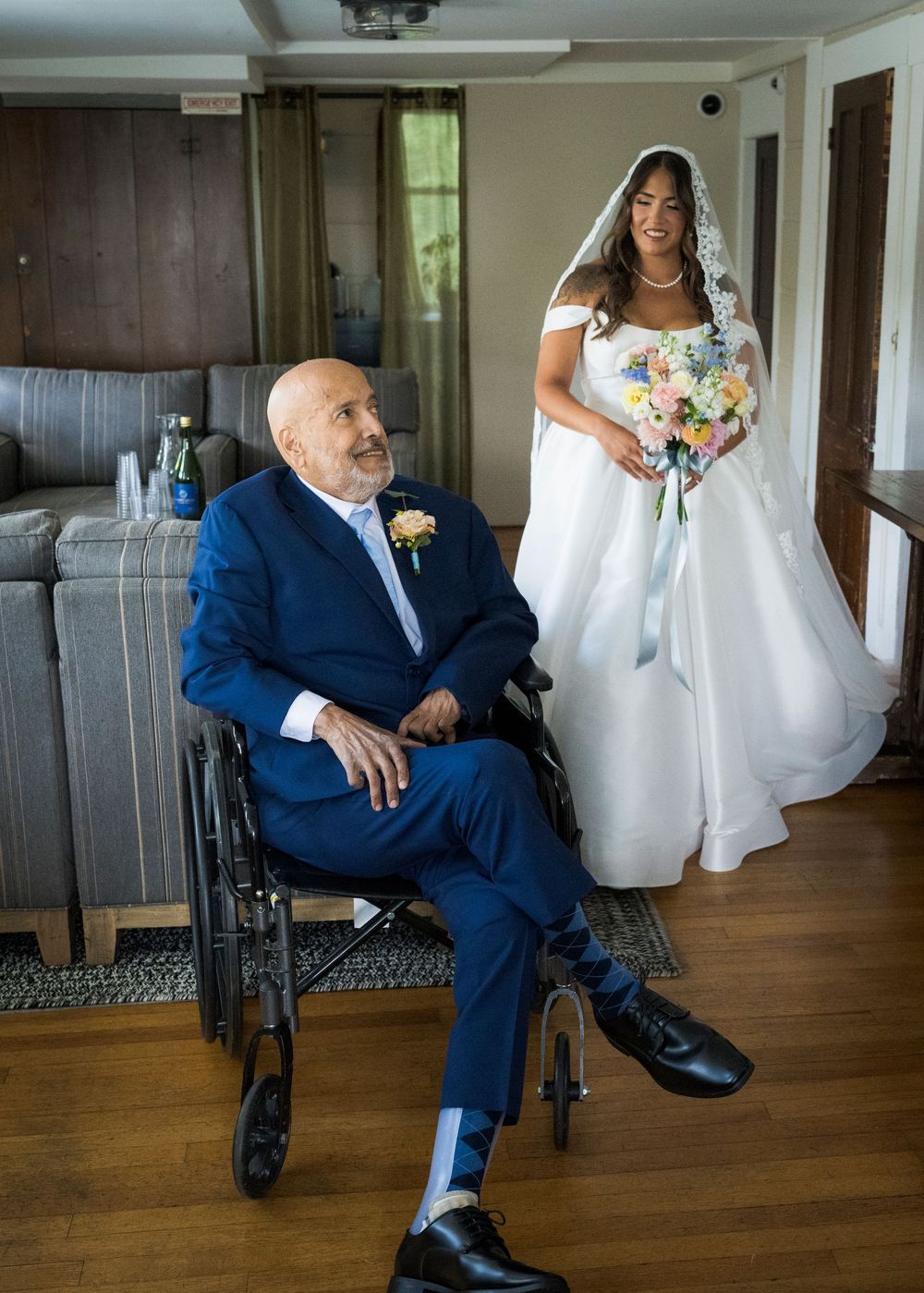 Bride in white gown holding bouquet stands with elderly man in wheelchair wearing blue suit at wedding.