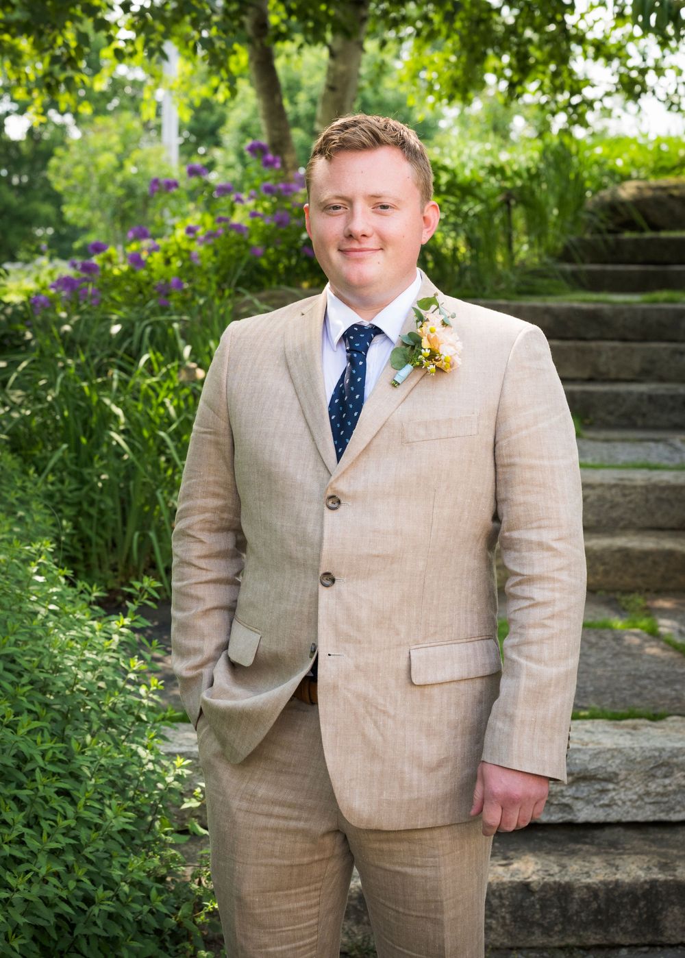 Smiling groom in tan linen suit with floral boutonniere posing in a garden.