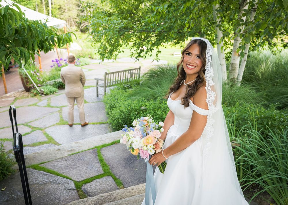 Smiling bride in white off-shoulder gown holding colorful bouquet during outdoor garden first look wedding moment.
