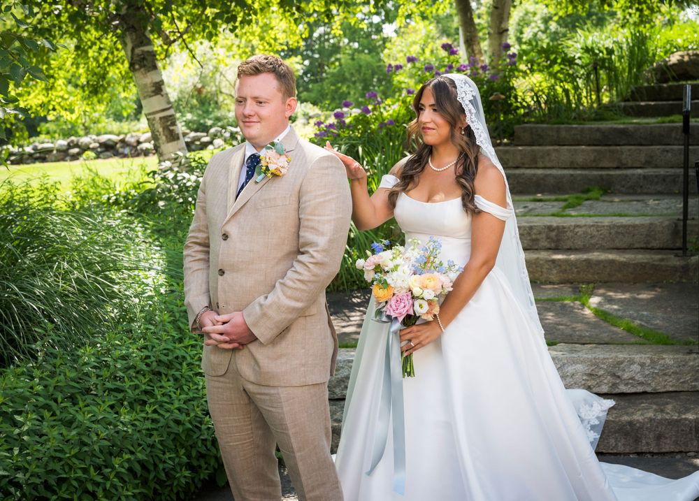 Bride in white gown holds bouquet beside groom in tan suit during outdoor first look wedding moment.