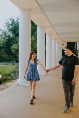 Couple holding hands walking under white columns colonnade, woman in blue floral mini dress smiling.