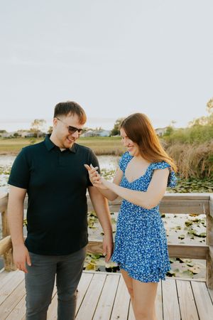 Happy couple on wooden dock admiring engagement ring, man in black polo and woman in blue floral dress.