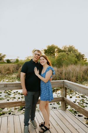 Couple posing on wooden boardwalk over lily pad pond at sunset, woman in blue floral dress showing engagement ring.