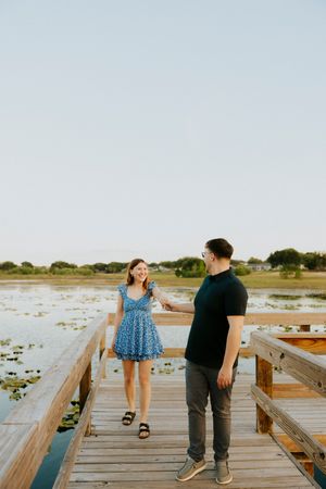 A smiling couple holds hands on a wooden boardwalk overlooking a serene lily pad-covered lake at sunset.