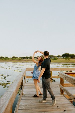 Couple dancing on a wooden dock overlooking a serene lake with lily pads at golden hour.