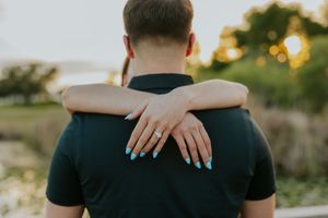 A woman with teal nails and engagement ring embraces a man from behind outdoors at sunset.