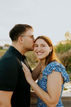 A man in a black shirt kisses a smiling woman in a blue floral dress on the cheek outdoors at golden hour.