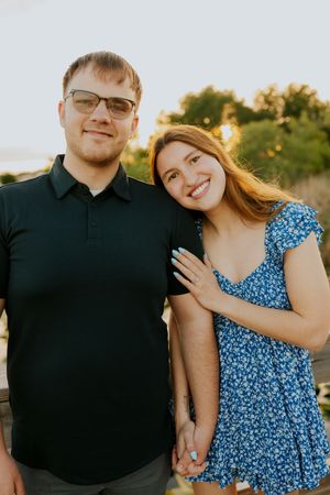 Smiling engaged couple holding hands on wooden boardwalk at sunset, woman in blue floral dress displaying ring.