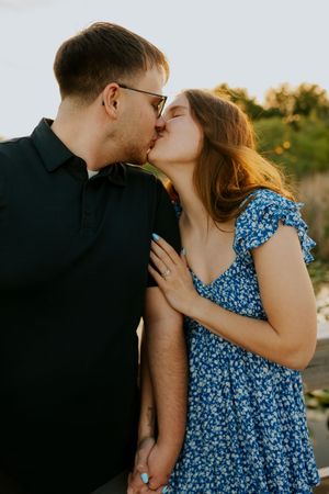 Couple sharing a romantic kiss outdoors at golden hour, woman in blue floral dress with engagement ring visible.