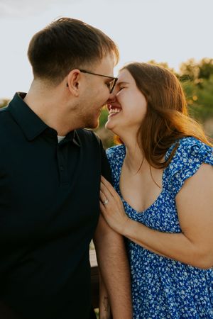 Joyful engaged couple laughing and touching noses at sunset, woman wearing blue floral dress.