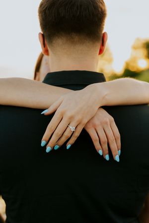 Close-up of a woman's hands with teal nails and diamond engagement ring wrapped around her partner.