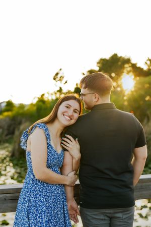Couple embracing at golden hour sunset, woman in blue floral dress smiling at camera on wooden bridge.