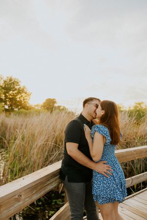Couple sharing a romantic kiss on a wooden boardwalk surrounded by golden tall grass at sunset.