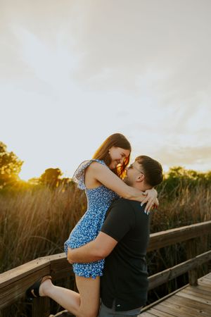 Couple embracing at golden sunset on wooden bridge, man lifting woman in blue floral dress.