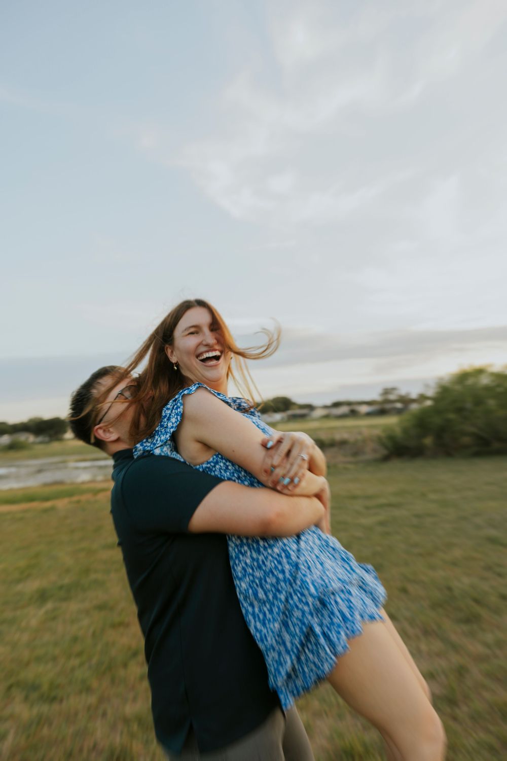 Joyful couple embracing outdoors at sunset, man in black shirt lifting laughing woman in blue floral dress in open field.