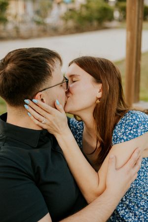 Couple sharing a romantic kiss outdoors, woman wearing blue floral dress and engagement ring, man in black shirt with glasses.