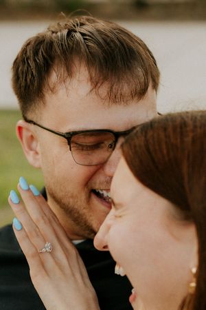 Couple laughing together closeup, woman showing diamond engagement ring with blue nails, man wearing glasses.