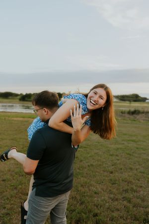 A joyful woman in a blue floral dress laughs as her partner lifts her in a sunny outdoor field near a lake.