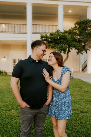 Couple smiling at each other on green lawn before white colonial building at golden hour.